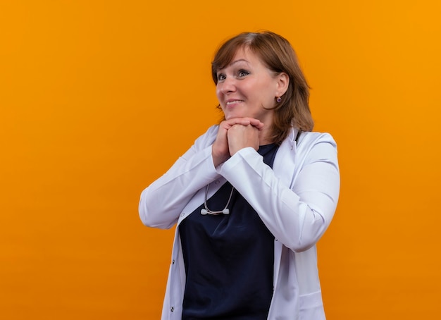 Excited middle-aged woman doctor wearing medical robe and stethoscope putting hands together on isolated orange wall with copy space