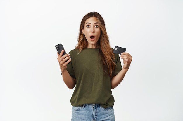 Excited middle aged woman looks happy, holding smartphone with credit card and stare amazed, buying online, shopping, buying on sale, standing against white background.