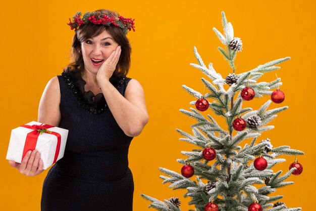 선물 패키지를 들고 장식 된 크리스마스 트리 근처에 서있는 목 주위에 크리스마스 머리 화환과 반짝이 갈 랜드를 입고 흥분된 중년 여성