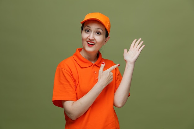 Excited middleaged delivery woman wearing uniform and cap looking at camera showing empty hand pointing at her hand isolated on olive green background