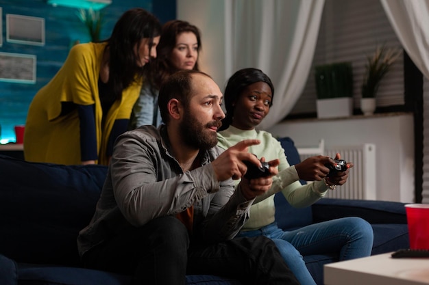 Excited multiethnic friends sitting on couch holding controller playing videogames during online competition enjoying spending time together. Group of people having fun relaxing in living room