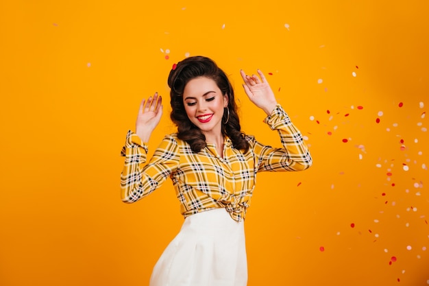 Excited pinup girl dancing and waving hands. Studio shot of blissful dark-haired woman in checkered shirt.