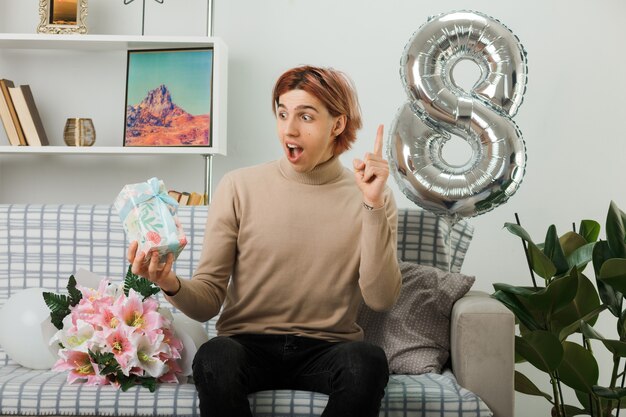 Excited points at up handsome guy on happy women day holding present sitting on sofa in living room