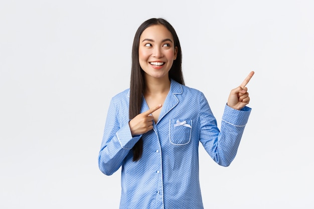 Excited pretty asian girl in blue pajamas pointing fingers upper right corner and looking interested. Pleased woman in jammies making choice, showing cool advertisement, white background