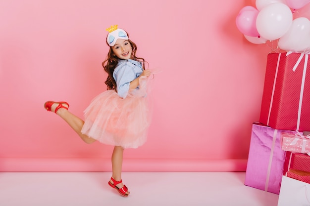 Excited pretty birthday girl with long brunette hair, in tulle skirt jumping, having fun isolated on pink background. Brightful celebration of amazing happy kid with giftboxes, balloons