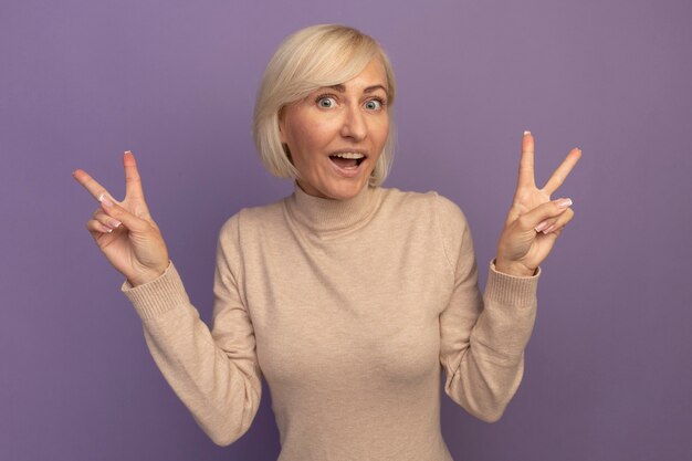 Возбужденная симпатичная славянская блондинка жестикулирует знак рукой победы двумя руками на фиолетовом
