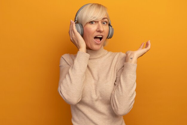 Excited pretty blonde slavic woman on headphones stands holding hand open on orange