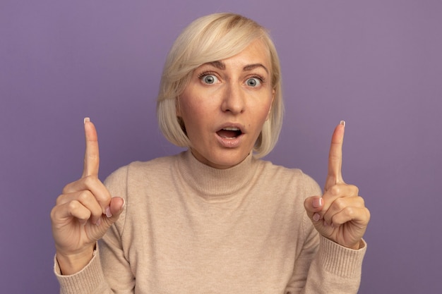 Excited pretty blonde slavic woman points up with two hands on purple