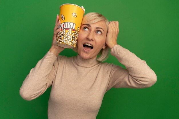 Возбужденная симпатичная славянская блондинка кладет руку на голову и держит ведро попкорна, глядя вверх изолированно на зеленой стене