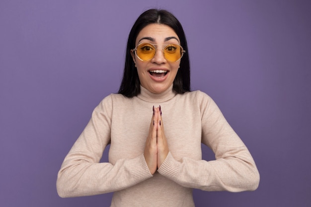 Excited pretty brunette caucasian girl in sun glasses holds hands together on purple