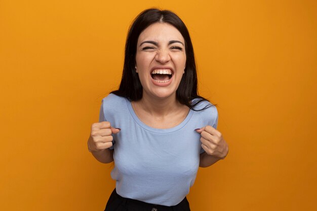 Excited pretty brunette woman keeps fists isolated on orange wall