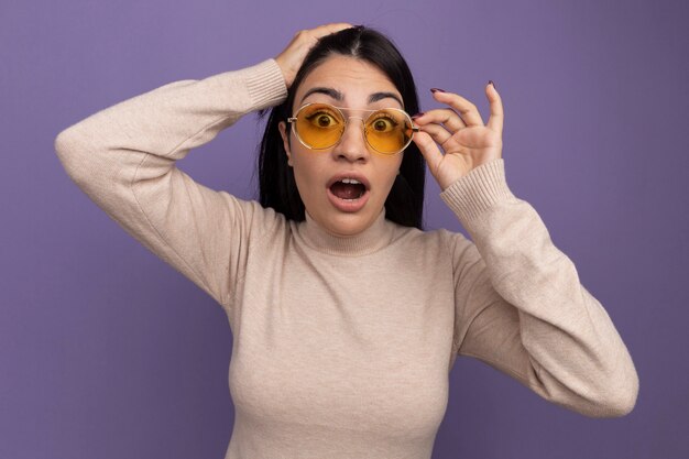 Free photo excited pretty brunette woman in sun glasses puts hand on head and looks at front isolated on purple wall