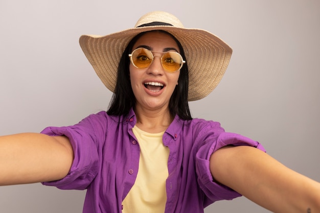 Excited pretty brunette woman in sun glasses with beach hat pretends to hold front taking selfie isolated on white wall