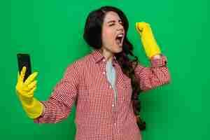 Free photo excited pretty caucasian cleaner woman with rubber gloves holding phone and raising fist up standing with closed eyes
