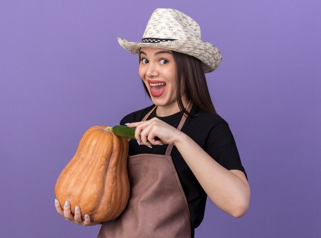 Excited pretty caucasian female gardener wearing gardening hat holding pumpkin and cucumber on purple