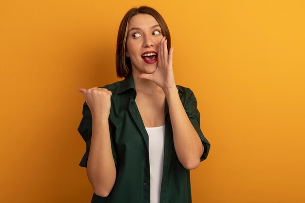 Excited pretty caucasian woman holds hand close to mouth looking and pointing at side on orange