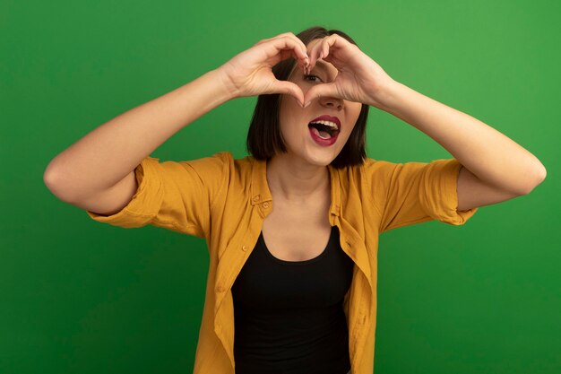 Excited pretty caucasian woman looks at camera through heart hand sign on green