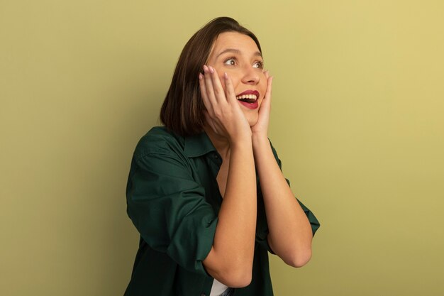 Excited pretty caucasian woman puts hands on face and looks at side on olive green