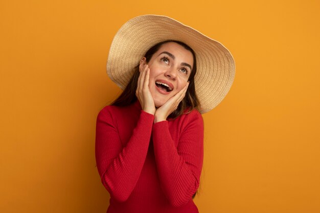 Excited pretty caucasian woman with beach hat  hands on face and looks up on orange