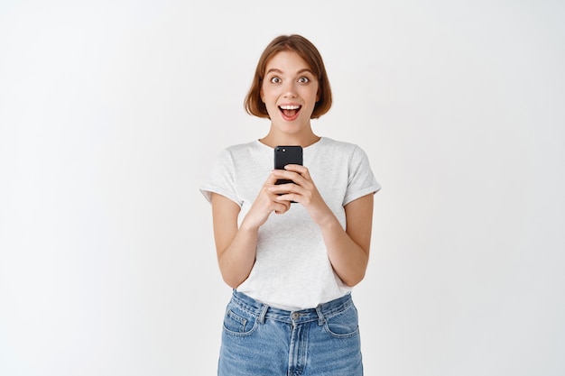 Excited pretty girl scream with joy after reading phone message, holding smartphone and looking amazed, checking out online promo, white wall