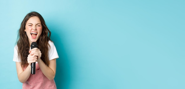 Foto gratuita bella ragazza eccitata che canta il karaoke tenendo il microfono e sorridendo felice in piedi sopra il backgrou blu