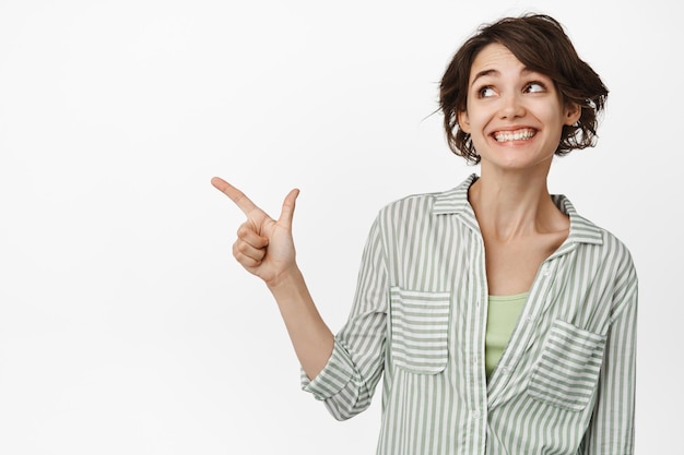Excited pretty millennial girl turn head and pointing thumb left, checking out good promo offer, demonstrating advertisement on copy space, white background.