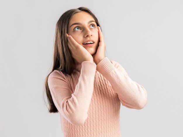 Excited pretty teenage girl looking up keeping hands on face isolated on white wall