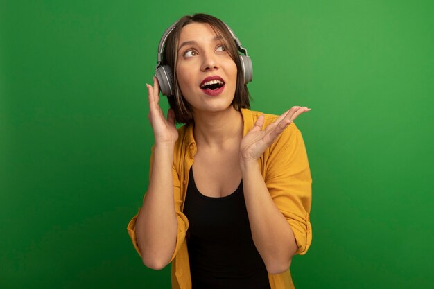 Excited pretty woman on headphones looks and points at side with hand isolated on green wall