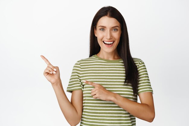 Excited pretty woman pointing fingers left smiling and showing store sale discount advertisement standing over white background