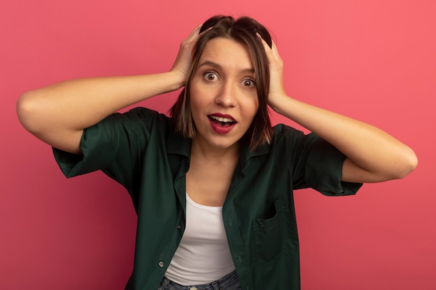 Excited pretty woman puts hands on head looking at front isolated on pink wall