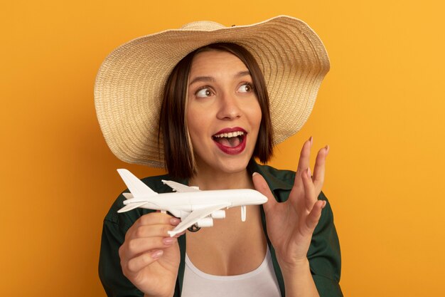 Excited pretty woman with beach hat holds model plane and looks at side isolated on orange wall