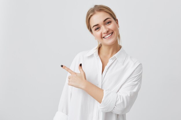 Excited pretty young blonde Caucasian woman wearing white shirt pointing her index finger sideways, smiling joyfully with her white teeth, showing something surprising on gray copy space wall