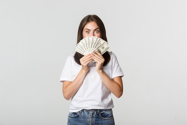 Excited pretty young woman holding money over face, standing white.