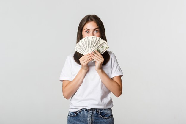 Возбужденная красивая молодая женщина, держащая деньги на лице, стоя белая.
