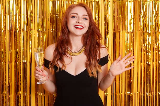 手を広げて、ワインのグラスを持って、新年を祝って、金色のきらめきで黄色の壁に立っている興奮した赤い髪の女性。