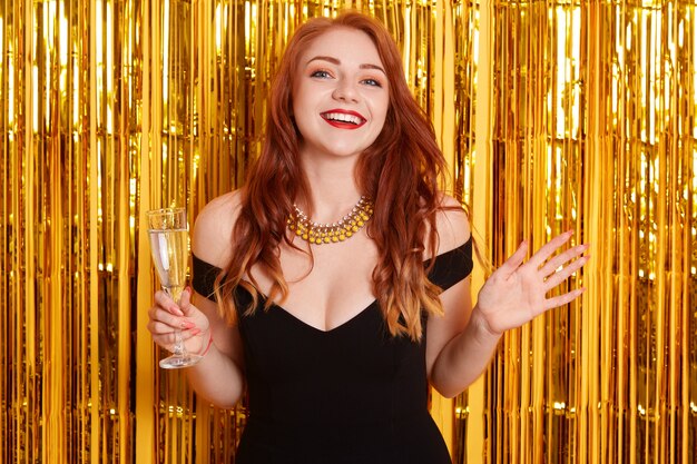 Excited red haired female with spreading hands aside, holding glass of wine, celebrating new year, standing against yellow wall with golden glitter.