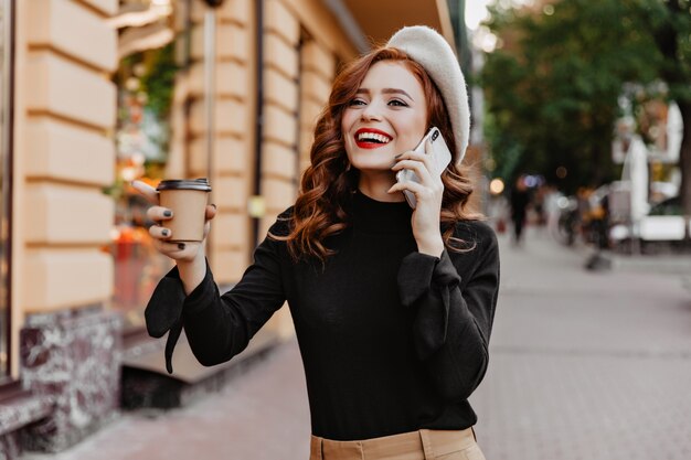 Excited red-haired girl drinking coffee on the street. Appealing stylish woman talking on phone on city wall.