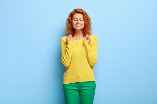 Excited red haired young woman makes wish, clenches fists and keeps eyes shut