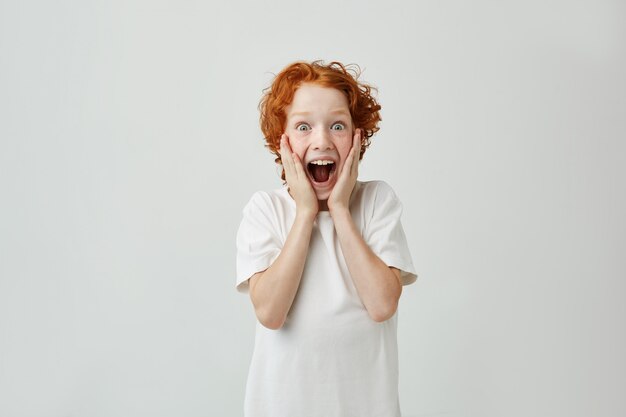 Excited redhead boy with freckles holding face with hands, with happy expression and opened mouth after parents gave him sweets.