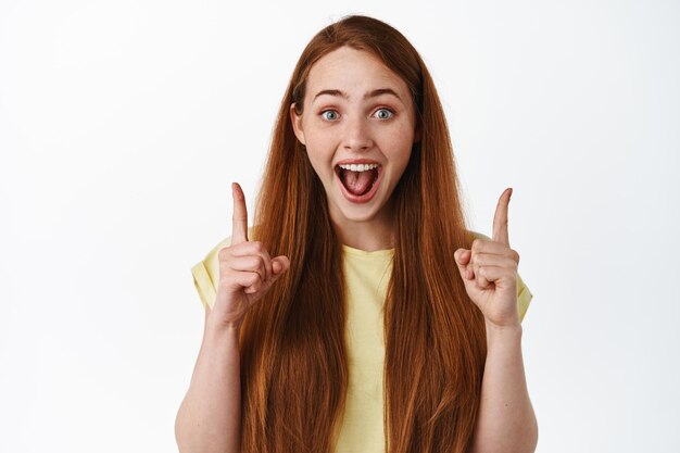 広告を表示し、面白がって笑顔で指を上に向けて、情報を提供し、何かクールな白い背景を発表する興奮した赤毛の女の子。