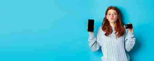 Free photo excited redhead girl showing mobile phone screen and credit card demonstrating online store or appli