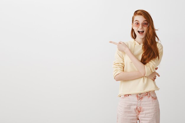 Excited redhead girl in sunglasses pointing finger left