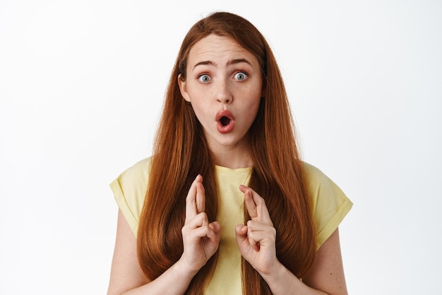 Excited redhead girl with crossed fingers gasping looking at results anticipating positive news and pray standing surprised against white background