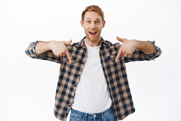 Excited redhead man pointing fingers down, showing awesome new promo offer, advertisement bellow, recommending website or store, standing over white wall