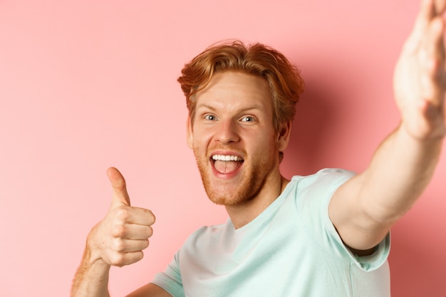 Excited redhead man tourist taking selfie and showing thumbs-up, holding camera with stretched out hand, standing over pink background