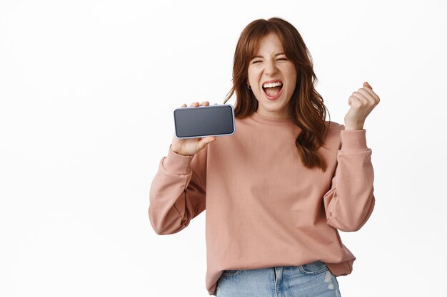 Возбужденная рыжая женщина показывает мобильный телефон с горизонтальным экраном, празднует и кричит от радости, показывает потрясающее приложение на смартфоне, стоя на белом фоне.