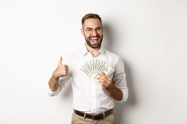 Excited rich man holding money, showing thumb up in approval, standing    