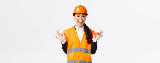 Взволнованная и довольная азиатская женщина-главный инженер, довольная удивительной работой, показывающая хороший жест, уверяет клиентов, что гарантирует идеальное качество, стоя в защитном шлеме на белом фоне