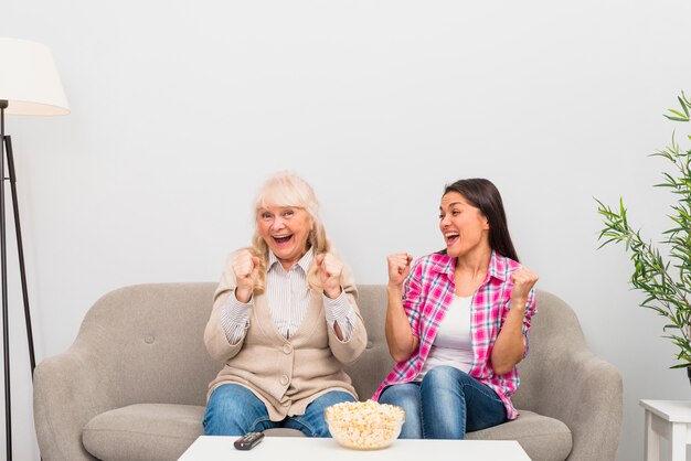 Excited senior mother and adult daughter sitting on sofa clenching their fist while watching television