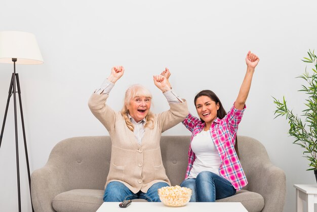 Возбужденные старшие мать и дочь, сидя на диване, поднимая руки во время просмотра телевизора